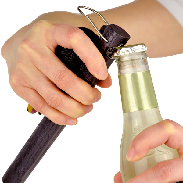 The Bear-B-Que Spatula is being shown with its bottle opener attachment, opening a gold colored glass bottle of soda.