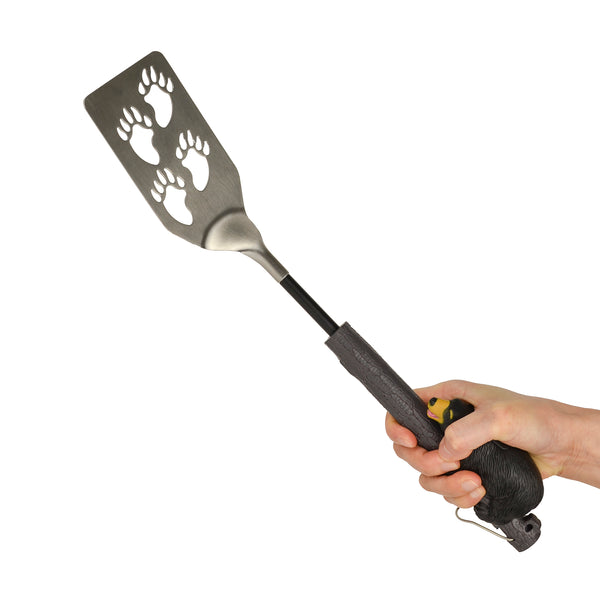 The Bear-B-Que Spatula is shown being held, with a large ergonomic handle, showing the handle resembling a brown bear with yellow markings climbing a tree. The spatula has bear paw cut outs in the spatula blade.